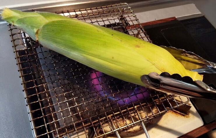 焼き網で焼いている皮つきトウモロコシ